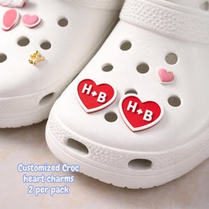 Puede incluir: Zapatos Crocs blancos con varios dijes decorativos. Se presentan dos dijes de corazón rojo con "H+B" en blanco. Otros dijes incluyen corazones rosas y una estrella dorada. El texto "Customized Croc heart charms 2 per pack" es visible.