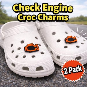 Puede incluir: Zuecos blancos con dijes en forma de motor naranja y negro. La imagen incluye el texto "Check Engine Croc Charms" en amarillo y una pegatina "2 Pack". Los zapatos están sobre una superficie de asfalto gris.