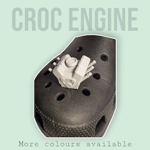 May include: A black Croc shoe with a small grey engine charm attached to it. The text "CROC ENGINE" is displayed above the shoe. The text "More colours available" is displayed below the shoe.