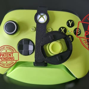 May include: A lime green Xbox controller with black accents and a black attachment. The controller has black buttons and a black joystick. The words "PATENT PENDING" are stamped on the controller in red.