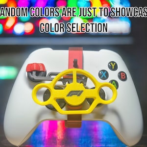 May include: A white Xbox controller with a yellow steering wheel attachment. The steering wheel has a black Formula 1 logo on it. The controller is sitting on a surface with a blurred background of colourful lights.
