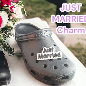 May include: A black Croc shoe with a white "Just Married" charm attached to the strap. The shoe is on a white surface with a bouquet of pink and red roses in the background.
