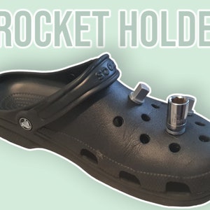 Könnte beinhalten: Ein schwarzer Crocs-Schuh mit zwei Metall-Steckschlüsselaufsätzen. Der Text "Crocket Holder" wird in hellgrüner Schrift über dem Schuh angezeigt. Der Schuh befindet sich auf einem hellgrünen Hintergrund.