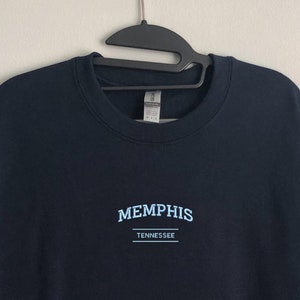Pu&ograve; includere: Una felpa blu navy con il testo "MEMPHIS" e "TENNESSEE" in lettere blu chiaro.