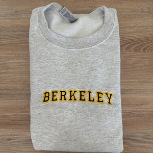 Może przedstawiać: Szara bluza z haftowanym logo "BERKELEY" w kolorze czarnym i żółtym.