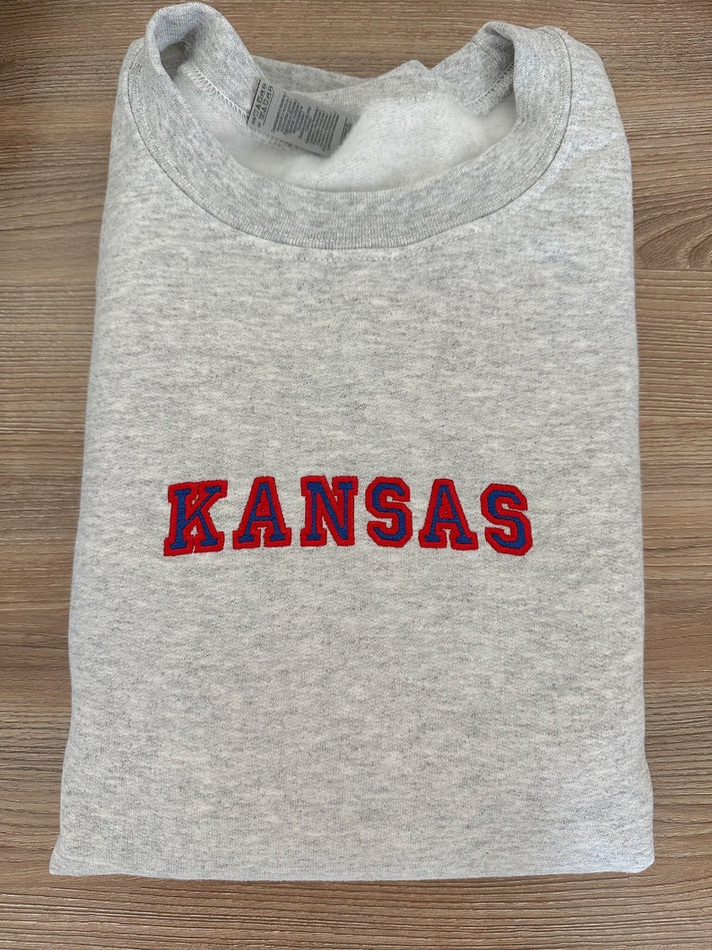 May include: Gray sweatshirt with the word "KANSAS" embroidered in red and blue lettering.