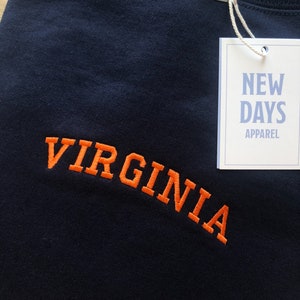 May include: A navy blue t-shirt with the word "VIRGINIA" embroidered in orange. The shirt has a tag attached that says "NEW DAYS APPAREL".