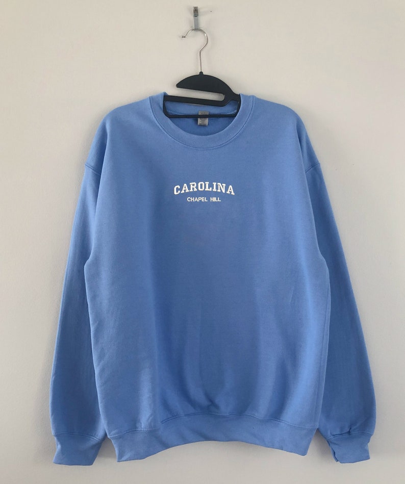 以下が含まれることがあります： 白文字で「CAROLINA」と「CHAPEL HILL」と書かれた、水色のスウェットシャツ。白い壁を背景に、黒いハンガーに吊るされています。