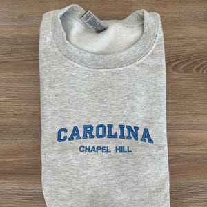 以下が含まれることがあります： 胸に青色の「CAROLINA」の文字が刺繍され、その下に「CHAPEL HILL」と刺繍された、折りたたまれたライトグレーのスウェットシャツ。スウェットシャツはクルーネックと長袖です。