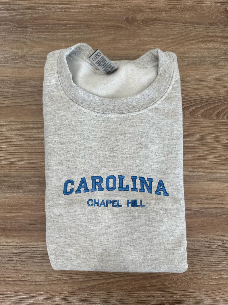 以下が含まれることがあります： 胸に青色の「CAROLINA」の文字が刺繍され、その下に「CHAPEL HILL」と刺繍された、折りたたまれたライトグレーのスウェットシャツ。スウェットシャツはクルーネックと長袖です。