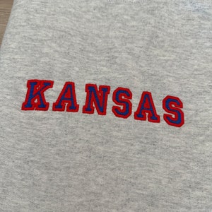 May include: Gray sweatshirt with the word "KANSAS" embroidered in red and blue lettering.