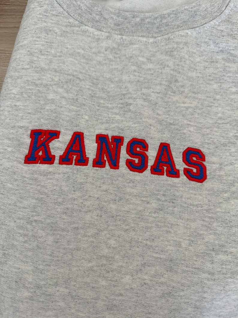 May include: Gray sweatshirt with the word "KANSAS" embroidered in red and blue lettering.