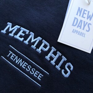 Pu&ograve; includere: Una felpa nera con testo ricamato in bianco che recita "MEMPHIS TENNESSEE" e un'etichetta bianca che recita "NEW DAYS APPAREL".