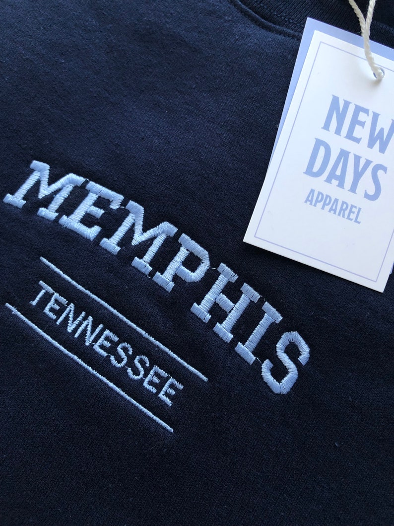 Pu&ograve; includere: Una felpa nera con testo ricamato in bianco che recita "MEMPHIS TENNESSEE" e un'etichetta bianca che recita "NEW DAYS APPAREL".