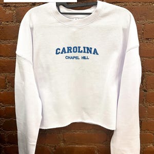以下が含まれることがあります： 「CAROLINA」の文字が青いブロック体で「CHAPEL HILL」の上に書かれた白いクロップドクルーネックのスウェットシャツ。画像の上部には、白い長方形の中に「CROPPED CREWNECKS AVAILABLE」というテキストがあります。スウェットシャツは黒いハンガーにかかっています。