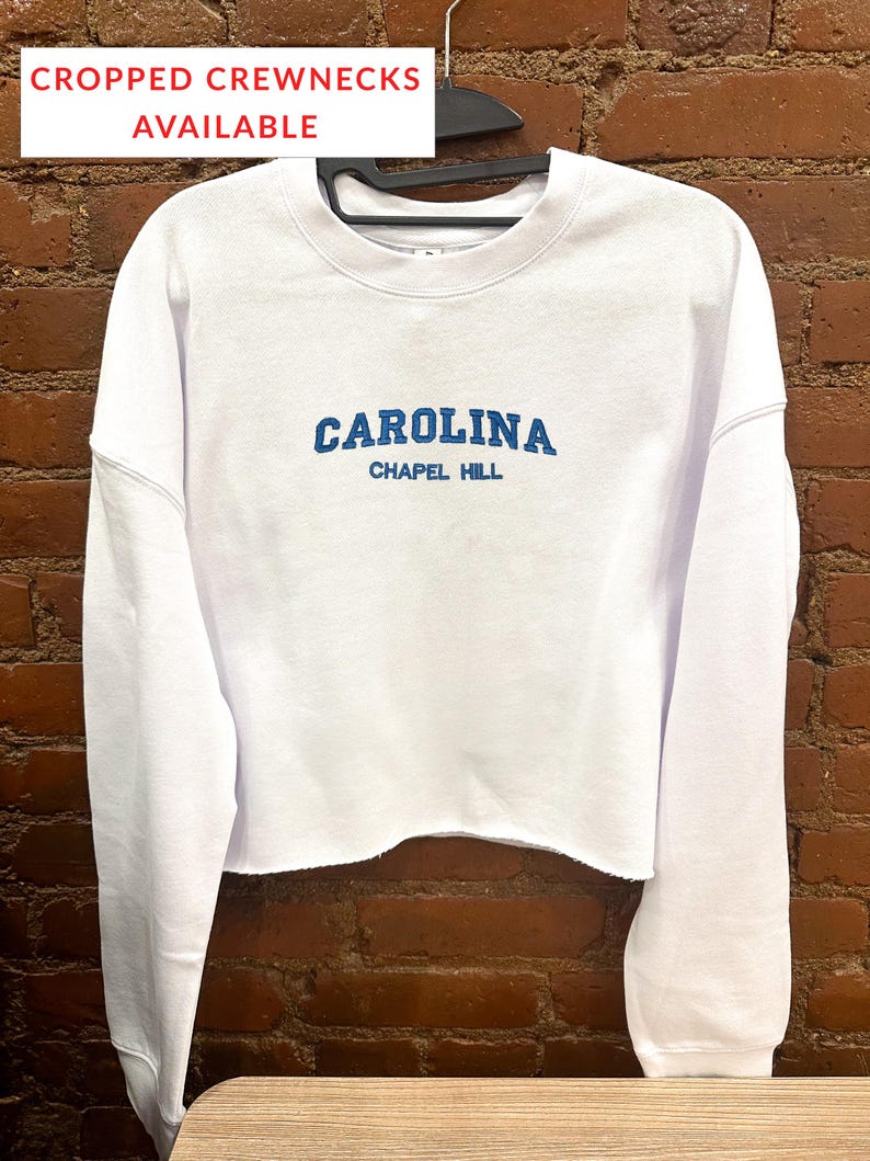 以下が含まれることがあります： 「CAROLINA」の文字が青いブロック体で「CHAPEL HILL」の上に書かれた白いクロップドクルーネックのスウェットシャツ。画像の上部には、白い長方形の中に「CROPPED CREWNECKS AVAILABLE」というテキストがあります。スウェットシャツは黒いハンガーにかかっています。