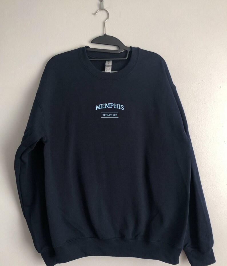 Pu&ograve; includere: Una felpa blu navy con il testo "MEMPHIS" e "TENNESSEE" in lettere blu chiaro.