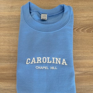以下が含まれることがあります： 白の「CAROLINA」の刺繍と、その下に「CHAPEL HILL」が入った水色のスウェットシャツ。 スウェットシャツは木製の表面にきちんと折りたたまれています。