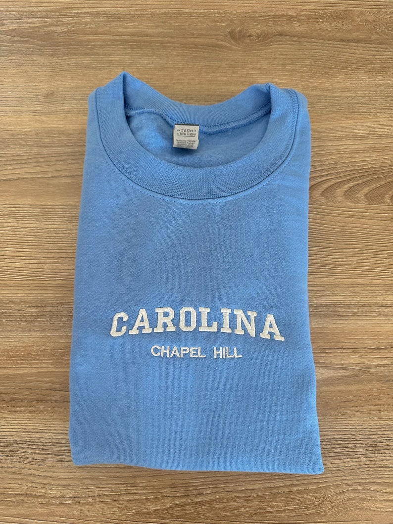 以下が含まれることがあります： 白の「CAROLINA」の刺繍と、その下に「CHAPEL HILL」が入った水色のスウェットシャツ。 スウェットシャツは木製の表面にきちんと折りたたまれています。