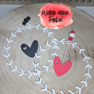 Könnte beinhalten: Zwei herzförmige Ornamente, eines schwarz und eines rot, hängen in weißen, blattförmigen Kränzen. Das rote Herz hat einen herzförmigen Ausschnitt. Der deutsche Text "Wähle deine Farbe!" ist auf rotem Aquarellhintergrund zu sehen.