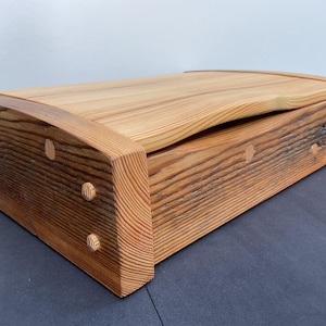 May include: A handcrafted wooden box with a natural finish. The box features a curved lid and is constructed with visible wood grain and dowel details. The warm tones of the wood create a rustic aesthetic. The box is approximately 46 cm long.