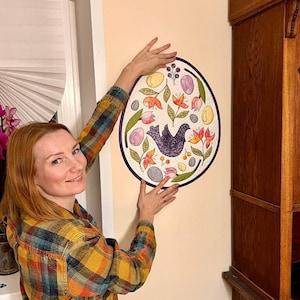 May include: A woman wearing a checked shirt is holding up a round, colourful wall hanging with a bird and floral design. The wall hanging is on a white wall.