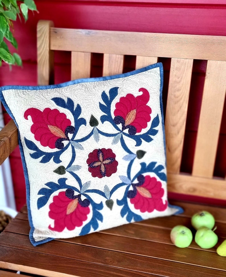 Peut inclure: Coussin carr&eacute; d&eacute;coratif avec un motif floral. Le coussin pr&eacute;sente un fond cr&egrave;me avec un motif de fleurs rouges, de feuilles bleues et une bordure bleue. Il repose sur un banc en bois avec des pommes vertes.