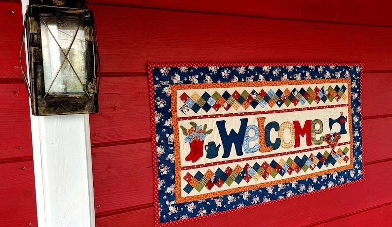 May include: A welcome sign made of patchwork fabric with a blue, red, and white color scheme. The sign says "Welcome" in large, colorful letters. The sign is hanging on a red wooden wall.
