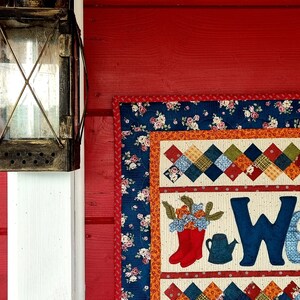 May include: A red wooden wall with a white post on the left side. A vintage lantern hangs on the post. A colorful quilted wall hanging with the word "Welcome" is on the right side of the wall.