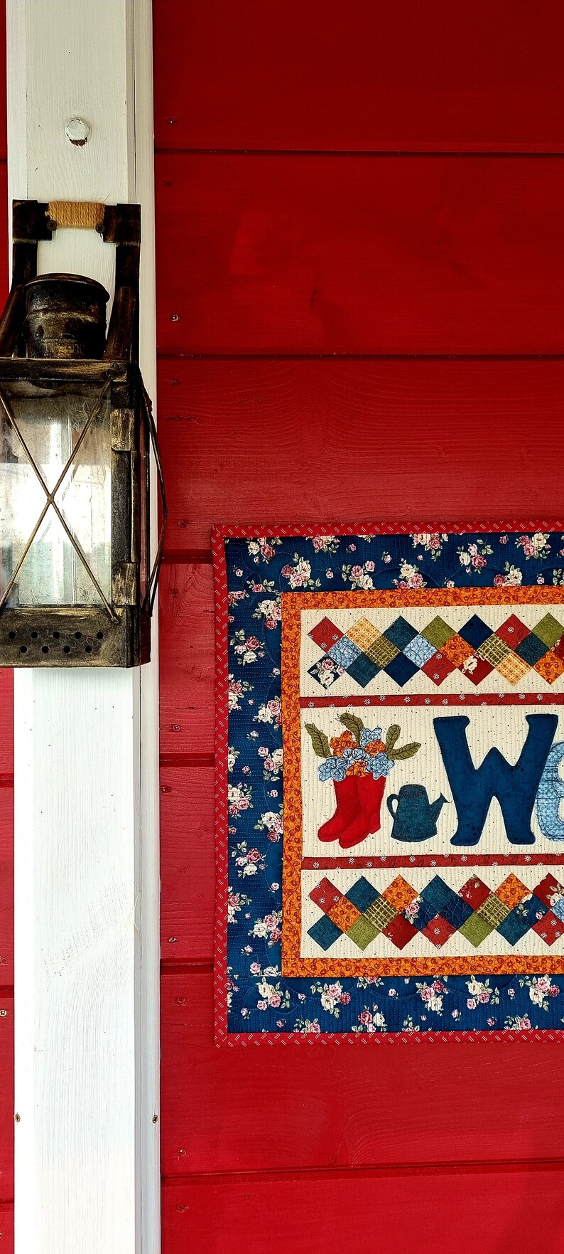 May include: A red wooden wall with a white post on the left side. A vintage lantern hangs on the post. A colorful quilted wall hanging with the word "Welcome" is on the right side of the wall.
