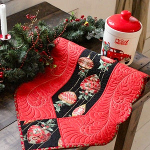 Pode incluir: Um corredor de mesa acolchoado vermelho e preto com uma borda festonada e um design de enfeites de Natal. O corredor está sobre uma mesa de madeira com uma guirlanda verde e um pote de cerâmica branco com uma tampa vermelha que diz "Feliz Natal".