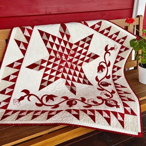 May include: A handmade quilt featuring a central red star design, framed by red and white geometric patterns and floral motifs. The quilt is displayed on a wooden bench, highlighting its detailed craftsmanship and design.