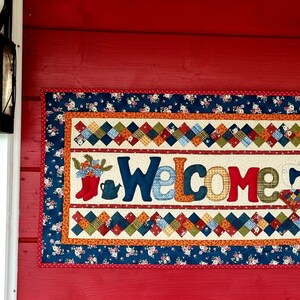 May include: A welcome sign with a patchwork design featuring a red boot, a watering can, and a sewing machine. The sign is framed with a blue and white floral border and hangs on a red wall.