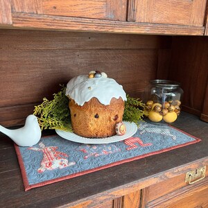 May include: A white and gold Easter cake with white icing and colourful sprinkles sits on a white plate with a blue and red bunny-themed table runner. The cake is on a wooden table with a wooden cabinet behind it.