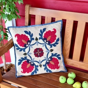 Peut inclure: Coussin carr&eacute; avec un fond cr&egrave;me et un motif floral rouge, bleu et marron. Il est pos&eacute; sur un banc en bois avec des pommes vertes. Le coussin a une bordure bleue et un motif matelass&eacute;.