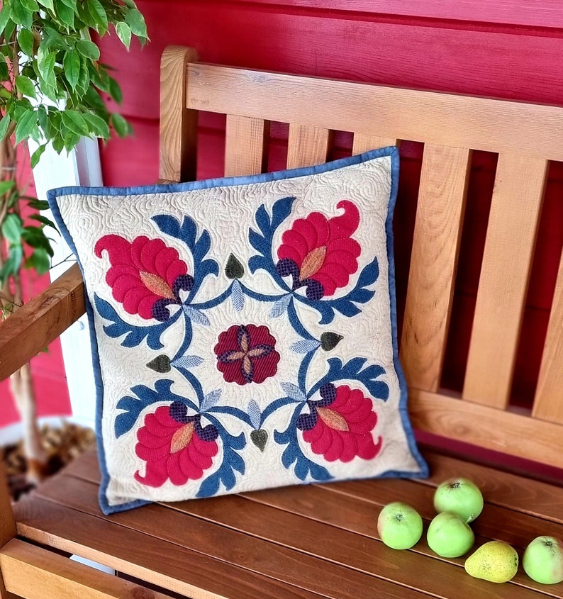 Peut inclure: Coussin carr&eacute; avec un fond cr&egrave;me et un motif floral rouge, bleu et marron. Il est pos&eacute; sur un banc en bois avec des pommes vertes. Le coussin a une bordure bleue et un motif matelass&eacute;.