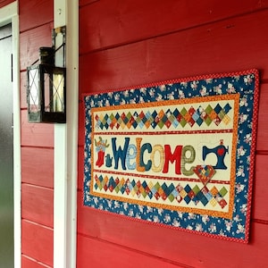 May include: A colorful welcome sign with a patchwork design and the word "Welcome" in large, colorful letters. The sign is hanging on a red wooden wall.