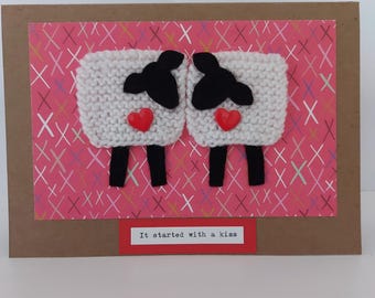 May include: A handmade greeting card featuring two white sheep with black faces and legs, each with a red heart. The sheep are on a pink background with white and pink crosses, set within a brown card. The text "It started with a kiss" is at the bottom.