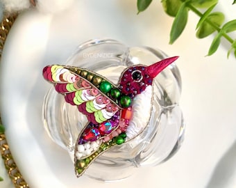 May include: A colorful hummingbird brooch made of sequins, beads, and embroidery. The hummingbird is in flight with its wings spread out. The brooch is on a white background with a gold chain border.
