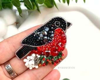 May include: A red and black beaded robin brooch with white beads and green accents. The brooch is held in a hand with a silver ring. The text "BYDENIZICE" is visible in the image.