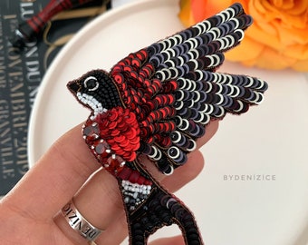 May include: A red, black, and white beaded bird brooch with a detailed design. The brooch is on a white plate and being held by a hand wearing a silver ring with the word "DENIZIC" engraved on it.