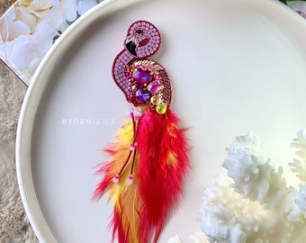 May include: A handcrafted flamingo brooch with a mosaic-style body, adorned with pink and purple crystals, and a beak. The brooch features vibrant red and yellow feathers. The text "BYDENIZICE" is visible.
