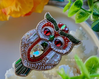 May include: A beaded owl brooch with red, green, and white accents. The owl has large, red eyes and a detailed design. The brooch is sitting on a white coral-like surface.