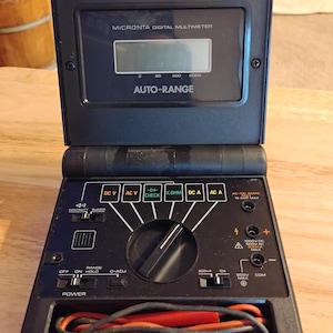 May include: Black Micronta digital multimeter with an open lid. The device features an LCD screen, various function buttons, and a dial. The text "MICRONTA DIGITAL MULTIMETER" and "AUTO-RANGE" are visible. Includes red and black test leads.