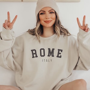 Può includere: Una felpa beige con la scritta "ROMA ITALIA" stampata sul davanti. La modella indossa un berretto beige e fa il segno della pace con entrambe le mani.
