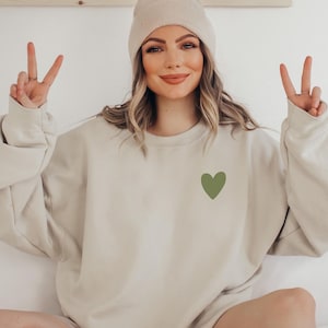 Op de afbeelding: Een vrouw draagt een beige sweatshirt met een groen hart op de voorkant. Ze draagt een witte beanie en heeft haar handen omhoog in een peace-teken.