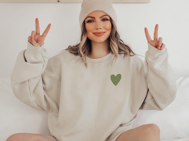 May include: A woman wearing a beige sweatshirt with a green heart on the front. She is wearing a white beanie and has her hands up in a peace sign.