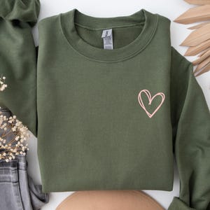 Puede incluir: Sudadera verde oliva con un contorno de corazón rosa en la parte delantera. La parte superior de manga larga está doblada, con una scrunchie a juego y unos vaqueros grises. Flores secas y una pieza decorativa beige también están en la imagen.