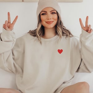 Puede incluir: Una mujer lleva una sudadera beige con un pequeño corazón rojo en la parte delantera. Está haciendo el signo de la paz con ambas manos. Lleva una boina beige claro.