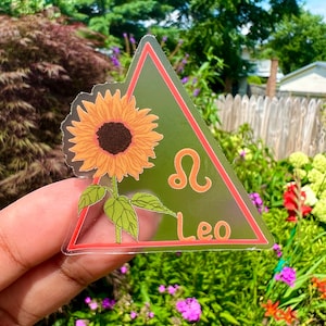 May include: A clear sticker with a red border in the shape of a triangle. The sticker features a sunflower with a yellow center and orange petals. The astrological symbol for Leo and the word "Leo" are printed in orange on the sticker.